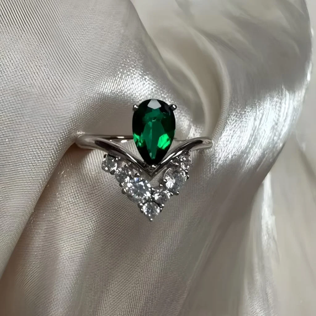 Lab-grown emerald pear-cut ring with V-shaped crown setting on white silk fabric, romantic wedding and engagement photography
