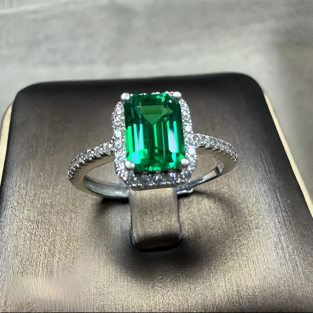 lab grown emerald halo ring S925 sterling silver on gray display surface emerald cut green gemstone baguette round diamond halo pavé band