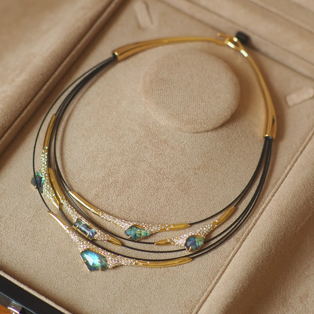 Multi-layer mother-of-pearl statement necklace close-up showing iridescent blue-green gemstones, pavé-set crystals, and gold-toned hammered metal bars on black leather cord, vintage artistic collar design by MAISON ELARA & CO