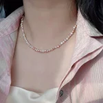 Model wearing natural freshwater baroque pearl necklace with pink collared shirt, showing multicolor pearls in peach, lavender, white, and rose gold tones at collarbone length with silver-toned clasp and heart-shaped extender charm by MAISON ELARA & CO