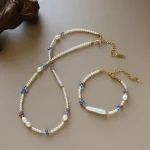 Dopamine freshwater pearl and colorful crystal beaded necklace and bracelet set flat lay on neutral surface, showing small round pearls, baroque pearl pendants, cobalt blue ruby red and soft pink crystal beads, and gold-toned clasp with extension chain by MAISON ELARA & CO