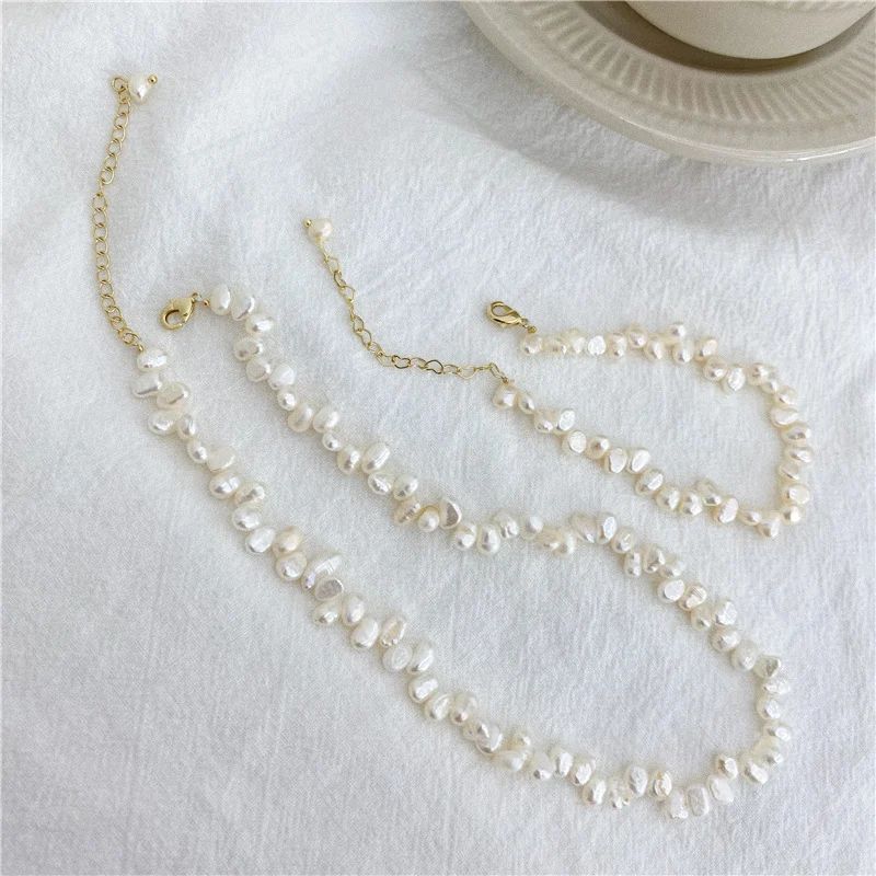 French baroque natural freshwater pearl necklace set of two pieces on white fabric surface with ceramic cup prop, showing irregular organic shaped pearls in ivory white cream tones with gold-toned lobster clasp and extender chain with pearl drop accent by MAISON ELARA & CO