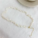 French baroque natural freshwater pearl necklace on white fabric surface with ceramic cup prop, showing irregular organic shaped pearls in ivory white cream tones with gold-toned lobster clasp and extender chain with pearl drop accent by MAISON ELARA & CO
