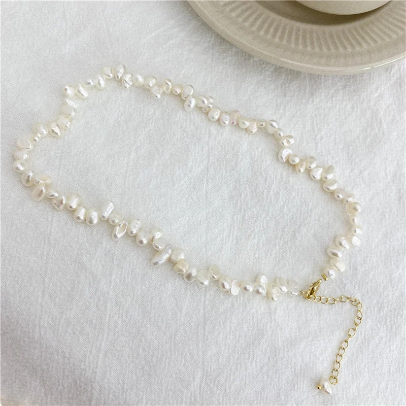 French baroque natural freshwater pearl necklace on white fabric surface with ceramic cup prop, showing irregular organic shaped pearls in ivory white cream tones with gold-toned lobster clasp and extender chain with pearl drop accent by MAISON ELARA & CO