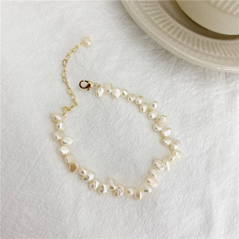 French baroque natural freshwater pearl bracelet and necklace on white fabric surface with ceramic plate prop, showing irregular organic shaped pearls in ivory white cream tones with gold-toned lobster clasp and extender chain by MAISON ELARA & CO