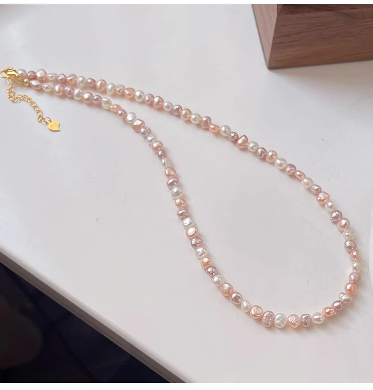 Natural freshwater baroque pearl necklace laid flat on light surface with gift box packaging, showing multicolor pearls in white, peach, lavender, and rose gold tones with silver-toned clasp and heart-shaped extender charm by MAISON ELARA & CO