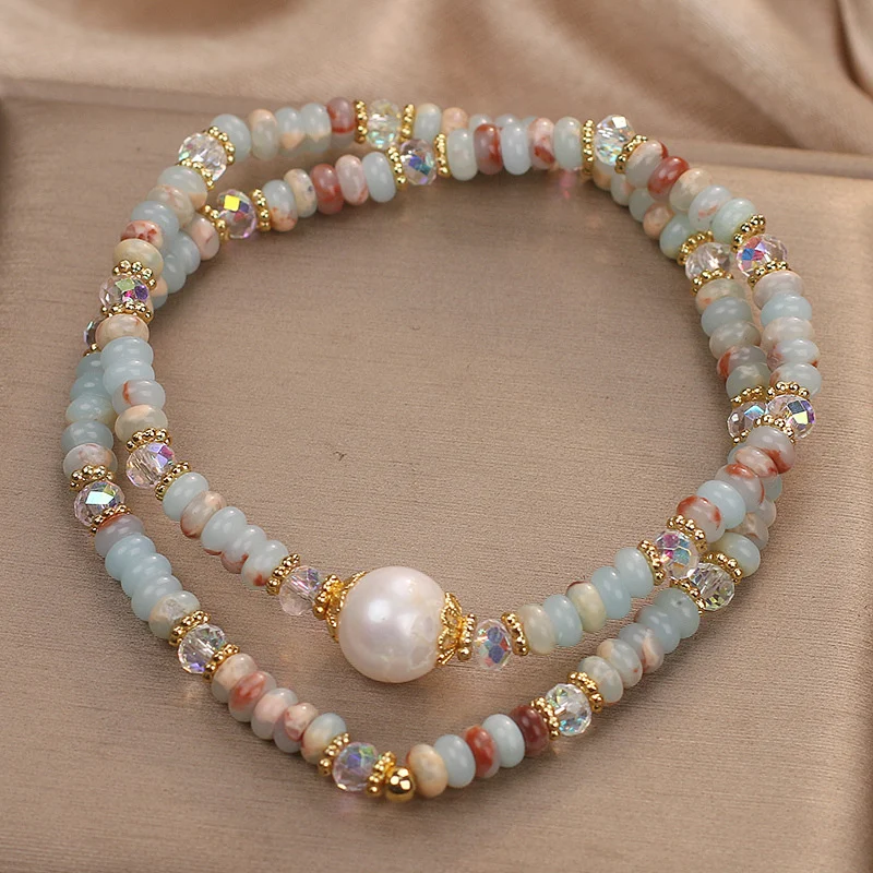 Top view of natural Shoushan stone pearl double-layer bracelet arranged in circle on beige satin, showing dual-strand elastic design with freshwater pearl focal stone and faceted AB crystal beads in blue white and rose tones