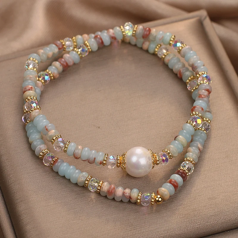 Overhead view of natural Shoushan stone double-layer bracelet with freshwater pearl centerpiece resting on folded beige satin gift box, dual-strand elastic design with AB crystal accents and gold-tone spacers