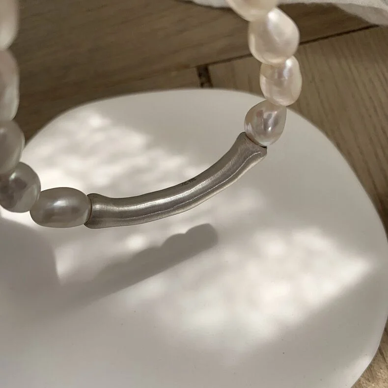 Close-up macro detail of 999 fine silver curved tube accent on baroque pearl bracelet showing brushed satin finish and organic curved shape on white display pedestal