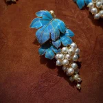 Close up of the blue enamel grape design with a faux pearl accent, showing gold tone metal detail on a deep brown background