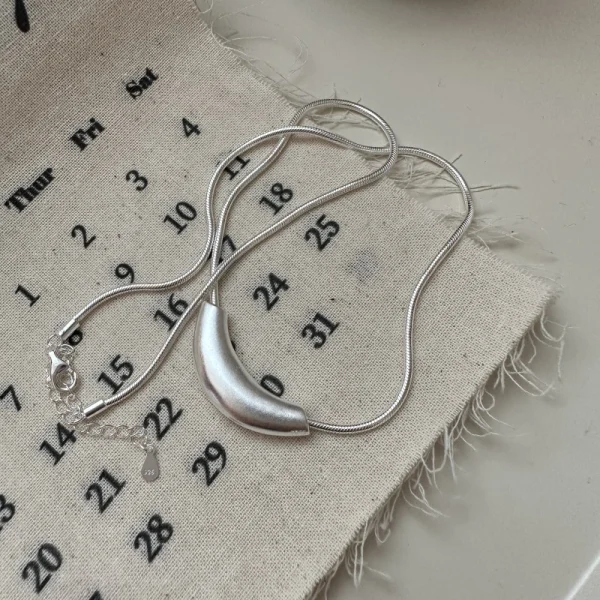S925 sterling silver matte curved crescent pendant snake chain necklace laid on linen calendar fabric