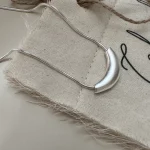 Close-up of matte curved crescent tube pendant S925 silver snake chain necklace on frayed linen fabric