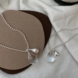 Two S925 sterling silver ball chain necklaces with teardrop crystal and silver pendants laid flat on linen and brown felt mat