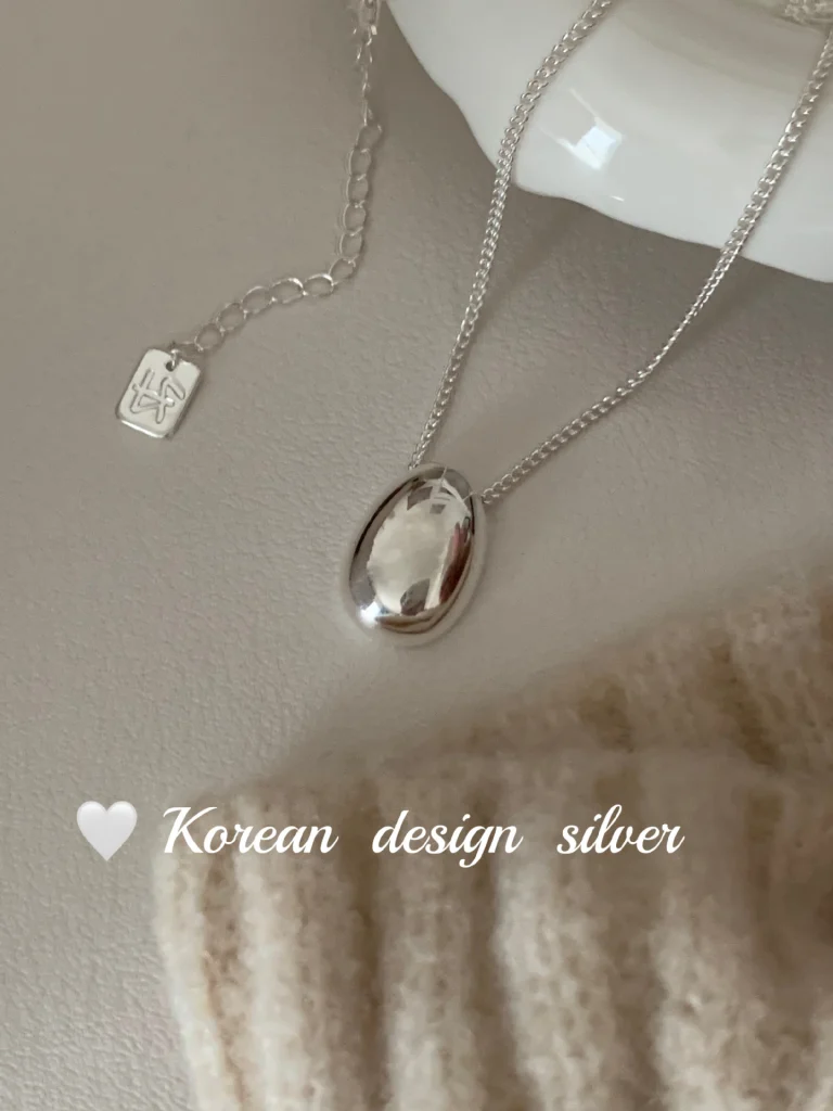 S925 sterling silver oval bean pendant necklace flat lay with cream knit fabric and white ceramic dish