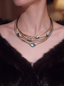 Multi-layer mother-of-pearl statement necklace worn on neck, featuring iridescent blue-green gemstones, pave-set crystals, and gold-toned metal bars on black leather cord, displayed on model in dark V-neck top