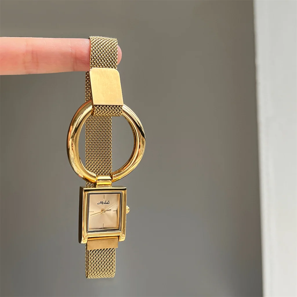 Close-up of the gold ring-detail square watch held between fingers, showing champagne dial