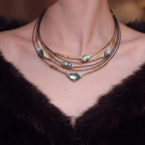 Multi-layer mother-of-pearl statement necklace worn on neck, featuring iridescent blue-green gemstones, pave-set crystals, and gold-toned metal bars on black leather cord, displayed on model in dark V-neck top