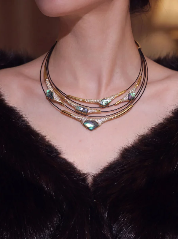 Multi-layer mother-of-pearl statement necklace worn on neck, featuring iridescent blue-green gemstones, pave-set crystals, and gold-toned metal bars on black leather cord, displayed on model in dark V-neck top