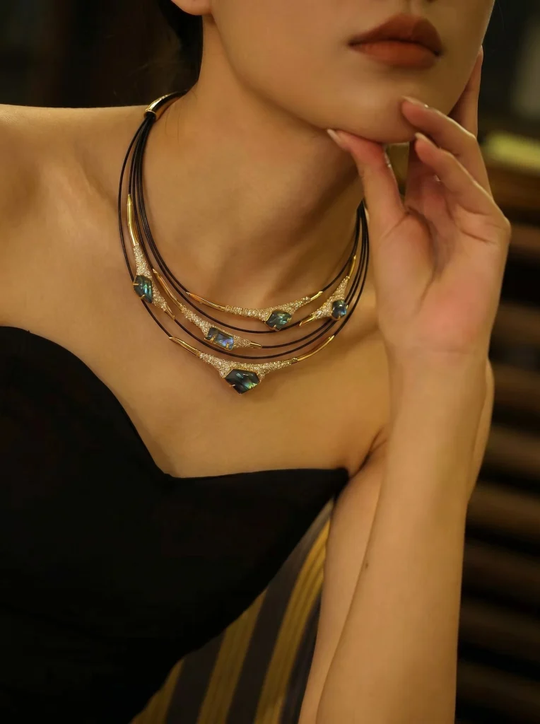 Model wearing multi-layer mother-of-pearl statement necklace with hand touching chin, wearing strapless black top, showing iridescent blue-green gemstones, pavé-set crystals, and gold-toned hammered metal bars on black leather cord under warm golden lighting by MAISON ELARA & CO