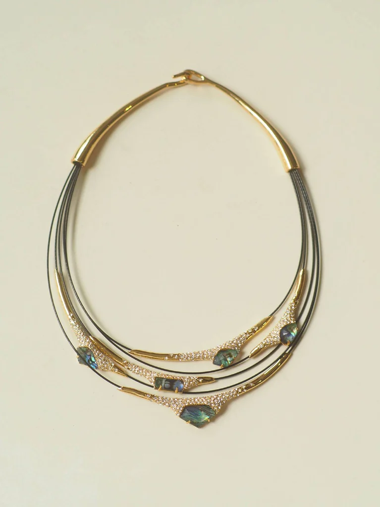 Multi-layer mother-of-pearl statement necklace laid flat showing full symmetrical design, four black leather cord strands, iridescent blue-green gemstones in teardrop and marquise shapes, pavé-set crystals, gold-toned metal bars with hammered texture, and hook clasp closure by MAISON ELARA & CO