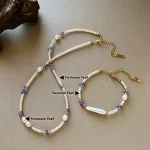 Dopamine freshwater pearl and colorful crystal beaded necklace and bracelet set laid flat on warm taupe surface, showing small round pearls, baroque pearl pendants, cobalt blue ruby red and soft pink crystal beads, and gold-toned clasp with extension chain by MAISON ELARA & CO